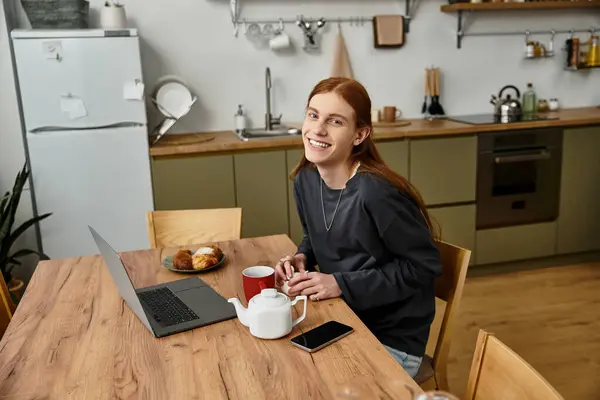 Relaxing at home, a young man with long hair sips tea by a laptop in his chic kitchen. — Stock Photo