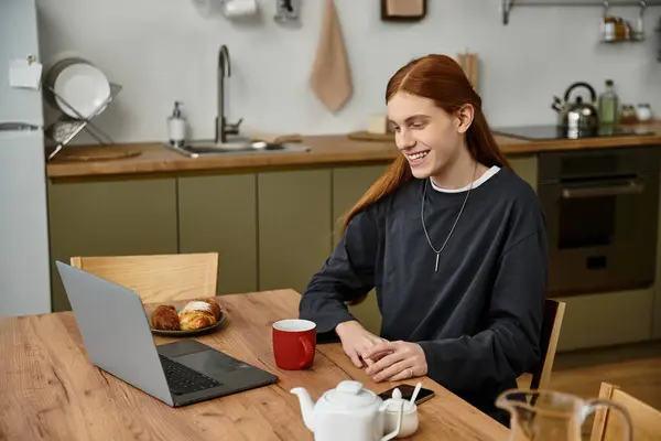 Young man with long hair smiles while enjoying breakfast and working on his laptop. — Stock Photo