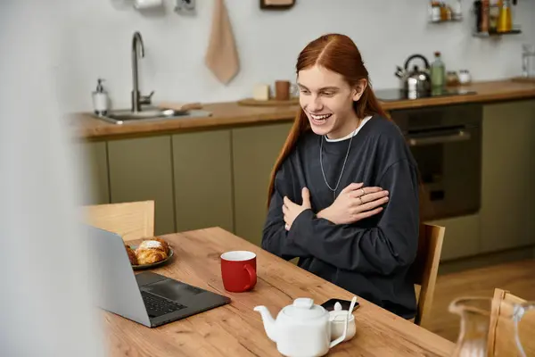 A young man with long hair sits at a modern kitchen table, smiling while enjoying his coffee. — Stock Photo