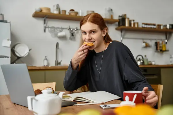 A stylish young man in casual clothes savors a pastry while journaling at home. — Stock Photo