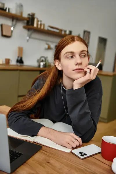 In un appartamento moderno, un giovane con i capelli lunghi si siede pensieroso ad una scrivania, rilassato. — Foto stock