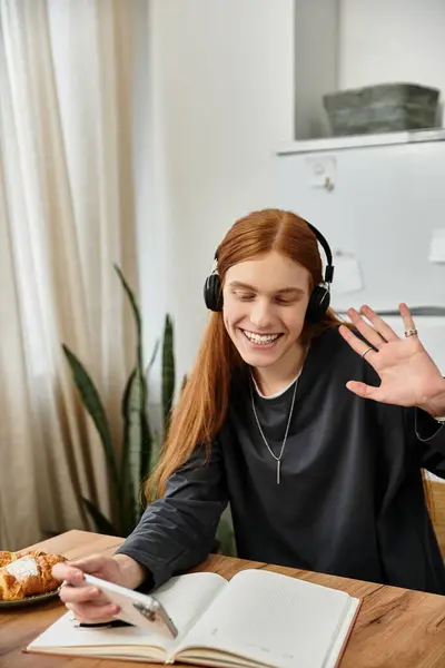 A young guy with long hair engages in a relaxed moment at home, smiling and waving. — Stock Photo