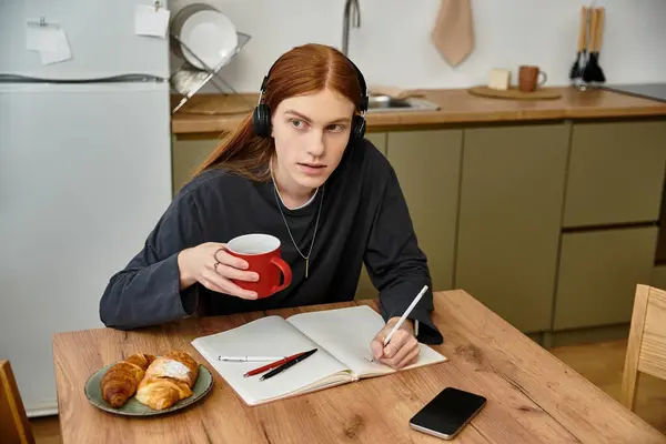 Un jeune adulte élégant aux cheveux longs se concentre sur ses études dans un appartement moderne, dégustant du café. — Photo de stock
