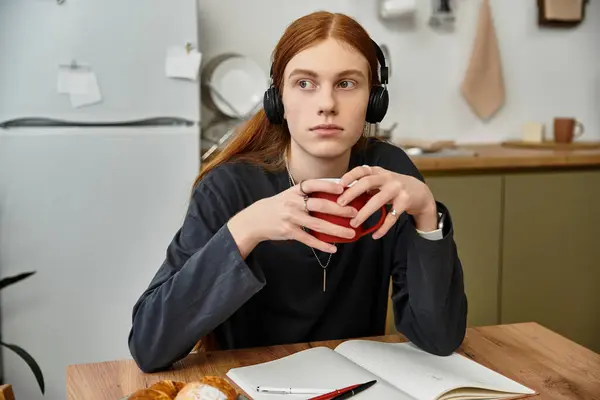 Un beau jeune homme aux cheveux longs se détend à la maison, sirotant une tasse vibrante tout en étudiant. — Photo de stock