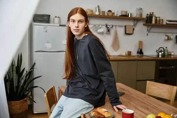 A laid-back young man with long hair enjoys breakfast at a wooden table in his apartment. — Stock Photo