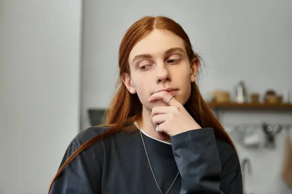 A young guy with long hair sits pensively in casual attire within his modern apartment. — Stock Photo