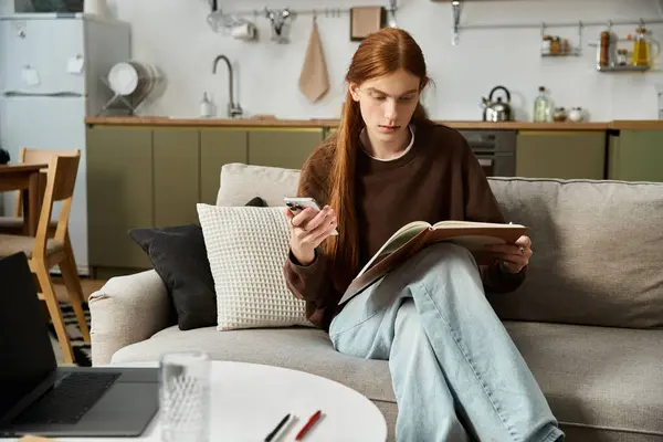 In a stylish apartment, a handsome young man relaxes on the couch, reading a book while texting. — Stock Photo