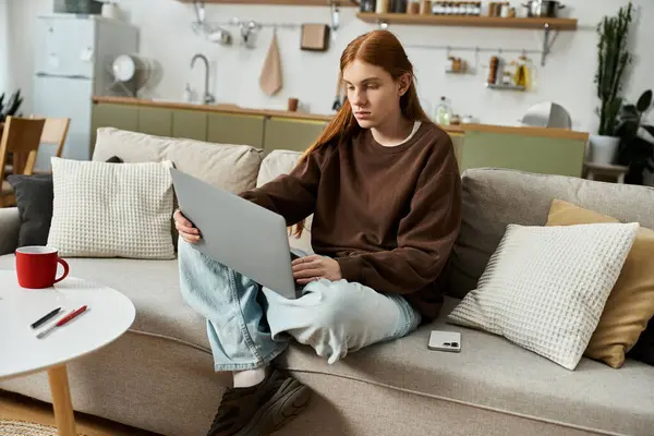 Young man with long red hair focuses on his laptop while lounging in a stylish living space. — Stock Photo