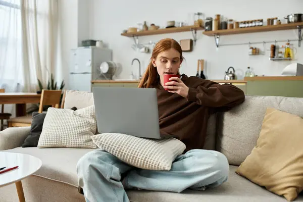 A young redhead man with long hair sits comfortably at home, sipping coffee while using a laptop. — Stock Photo