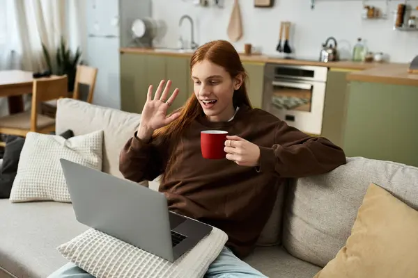 A young man with long red hair sips from a red cup during a video call in his stylish home. — Stock Photo
