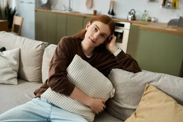 A young man with long red hair relaxes at home in a cozy sweatshirt, embracing a pillow. — Stock Photo