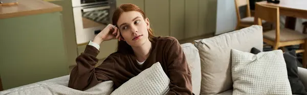 A young redhead in a cozy sweatshirt relaxes thoughtfully on a sofa in his apartment, banner — Stock Photo