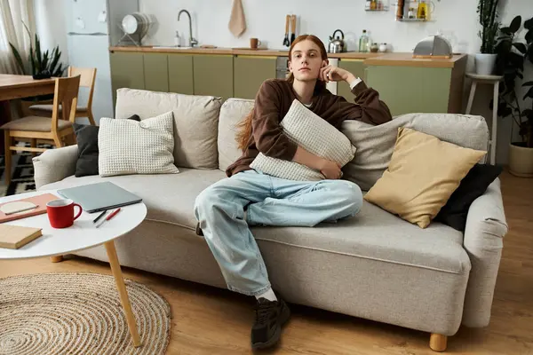A stylish young man with long red hair sits on a couch, deep in thought at home. — Stock Photo