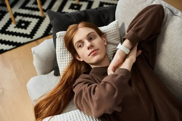 A handsome young man with long red hair lounges comfortably at home, enjoying leisurely moments. — Stock Photo