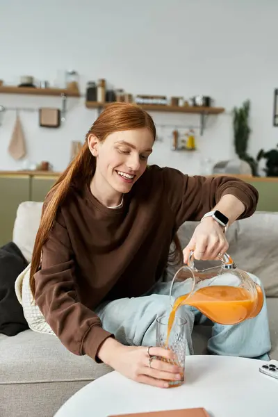 Un joven alegre con el pelo largo y rojo vierte jugo en un vaso mientras se relaja en el interior. - foto de stock