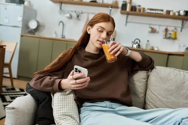 Ein junger Rotschopf entspannt sich in seiner stilvollen Wohnung, nippt an einem Drink und benutzt sein Handy. — Stockfoto