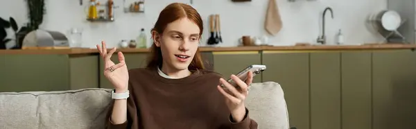 A stylish redheaded young man relaxes at home, engaged in a lively conversation on his phone, banner — Stock Photo