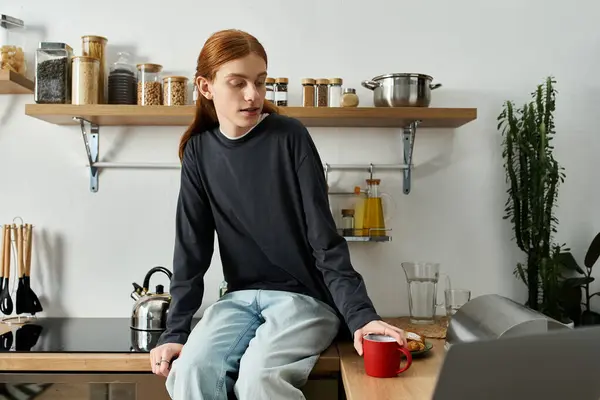 Young man with long red hair sits on a countertop, enjoying a warm drink in a stylish apartment. — Stock Photo