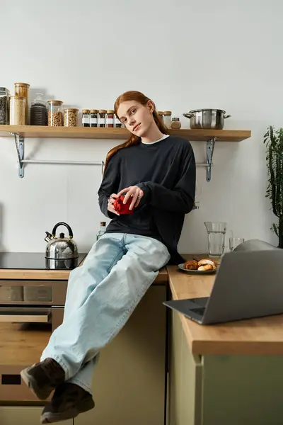 A young man with long red hair relaxes in his trendy kitchen while enjoying a drink. — Stock Photo
