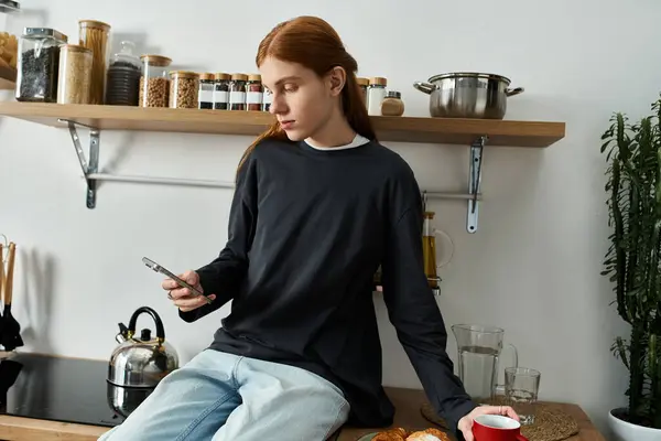 A good looking young man with long red hair relaxes in his stylish kitchen while checking his phone. — Stock Photo