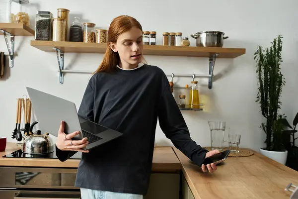 A young guy with long red hair balances his laptop and phone in a chic kitchen. — Stock Photo