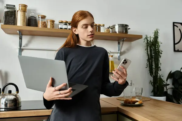 Charming young guy with long red hair balances laptop and phone in modern apartment. — Stock Photo