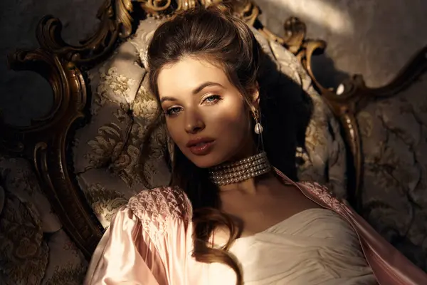 A beautiful young woman rests elegantly on an ornate chair, bathed in soft sunlight. — Stock Photo
