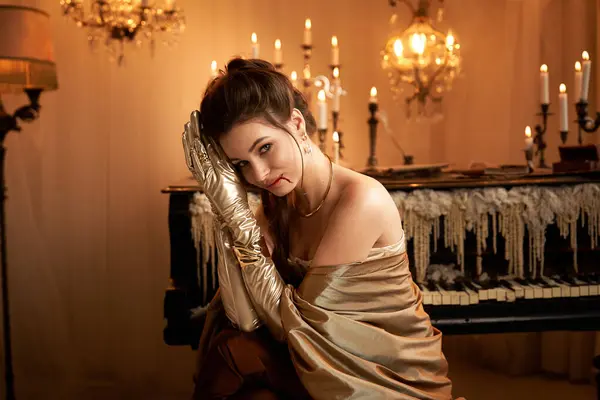 A young woman in elegant gold leans against a piano, surrounded by flickering candles. — Stock Photo