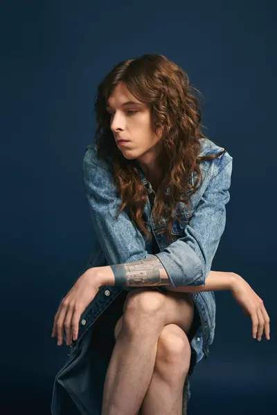 Confident young man with curly hair sits thoughtfully against a deep blue backdrop. — Stock Photo