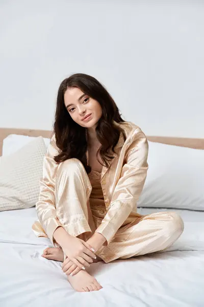 A beautiful young woman sits cross legged on her bed in golden loungewear, radiating calm. — Stock Photo