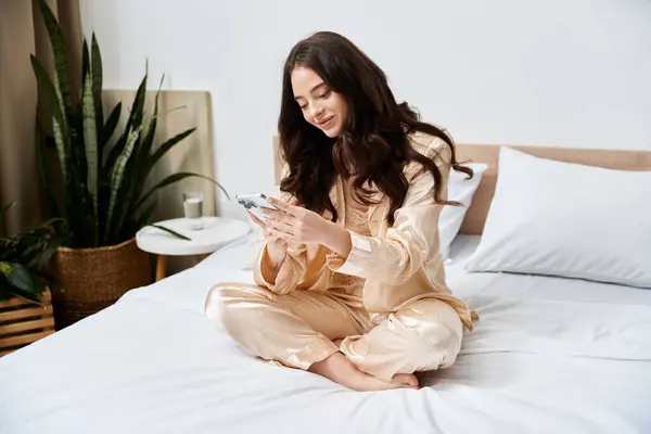 A beautiful young woman sits comfortably on her bed, smiling at her phone in soft pajamas. — Stock Photo