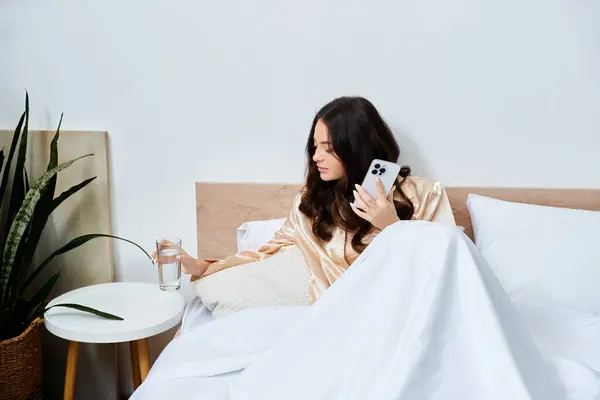 Rilassandosi a letto, una bella giovane donna si gode un bicchiere d'acqua mentre usa il suo smartphone. — Foto stock