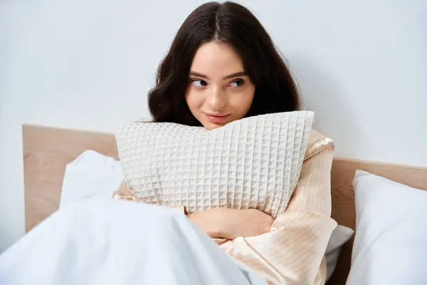 A beautiful young woman sits in bed, wrapping her arms around a pillow, smiling with joy. — Stock Photo