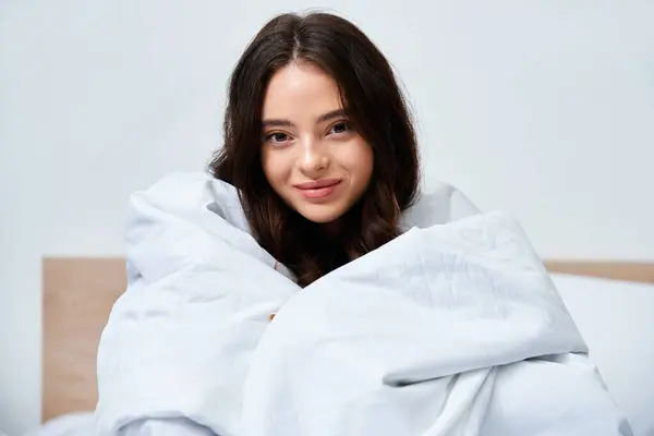 A young woman sits on a bed, wrapped in a soft blanket, smiling with a charming expression. — Stock Photo