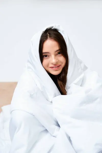 Une belle jeune femme sourit doucement tout en étant drapée dans un tissu blanc doux et confortable à l'intérieur. — Photo de stock