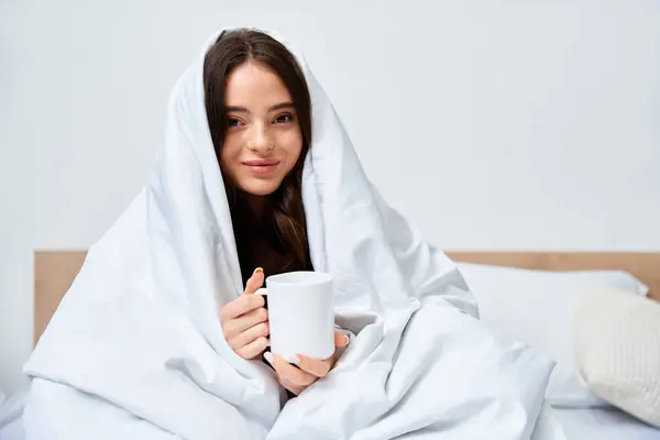 A beautiful young woman enjoys a warm drink while snuggled in a soft blanket in her cozy bedroom. — Stock Photo