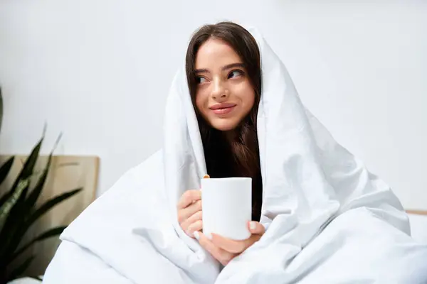 A young woman relaxes on a cozy bed, wrapped in a soft blanket, sipping a warm drink. — Stock Photo
