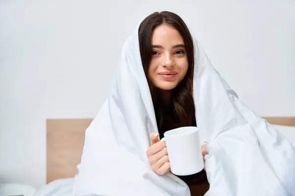 A beautiful young woman enjoys a hot beverage while wrapped in a soft blanket in her cozy room. — Stock Photo