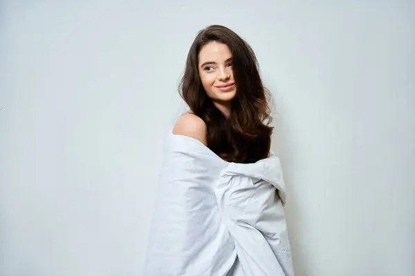 Une belle jeune femme aux cheveux fluides sourit doucement enveloppée dans une couverture confortable. — Photo de stock