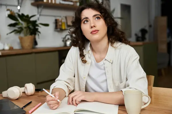 A young woman with curly hair contemplates as she writes notes at her home work desk. — Stock Photo