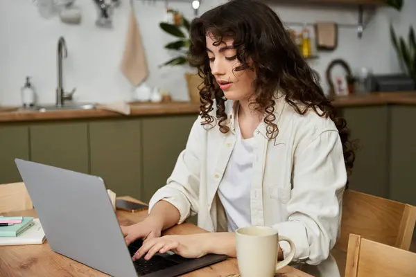 A young woman types intently on her laptop at home, savoring her coffee. — Stock Photo