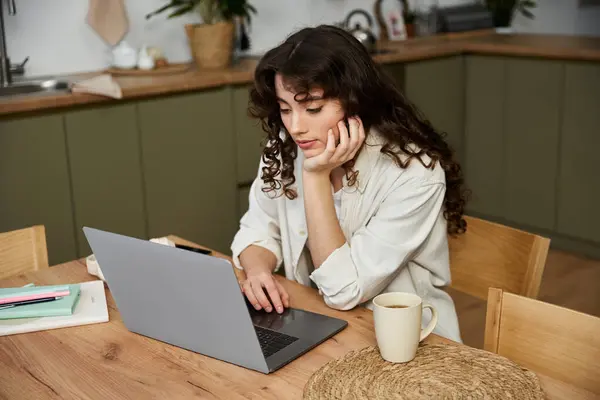 Una giovane donna con i capelli ricci si concentra intensamente sul suo computer portatile sorseggiando caffè a casa. — Foto stock