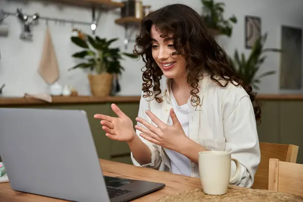 Sorridente giovane donna interagisce eccitatamente durante la sua videochiamata a casa con una bevanda calda nelle vicinanze. — Foto stock