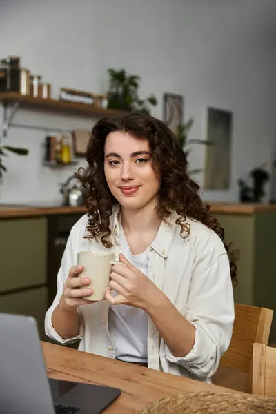 A young woman with curly hair sits at a wooden table, sipping a warm beverage and smiling contently. — Stock Photo