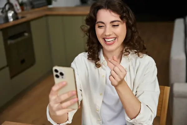 A cheerful young woman smiles while engaging with friends on her smartphone in a cozy home setting. — Stock Photo