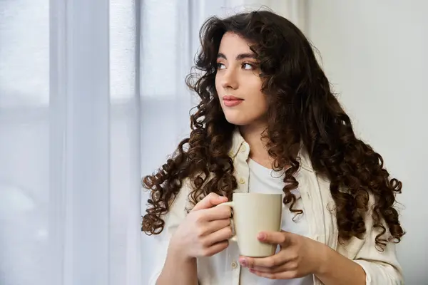 A young woman sits alone at home, sipping her warm drink and gazing outside. — Stock Photo