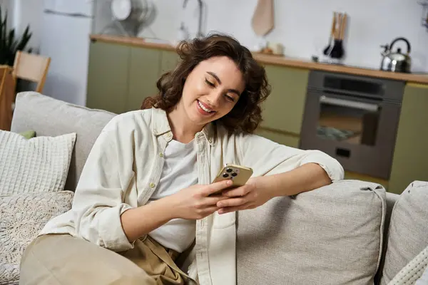 Una hermosa joven con el pelo rizado sonríe mientras usa su teléfono inteligente en un acogedor espacio de vida. - foto de stock