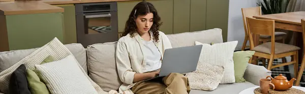 Young woman sits comfortably on a couch, working on her laptop in a warm, inviting home setting. — Stock Photo