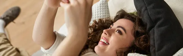 A beautiful woman sprawls on her couch, laughing and fully engaged in her mobile game at home. — Stock Photo