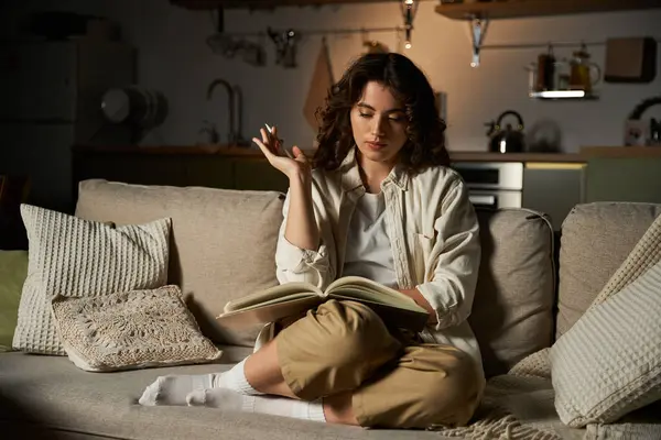 A beautiful young woman enjoys a quiet evening at home, absorbed in reading on her couch. — Stock Photo